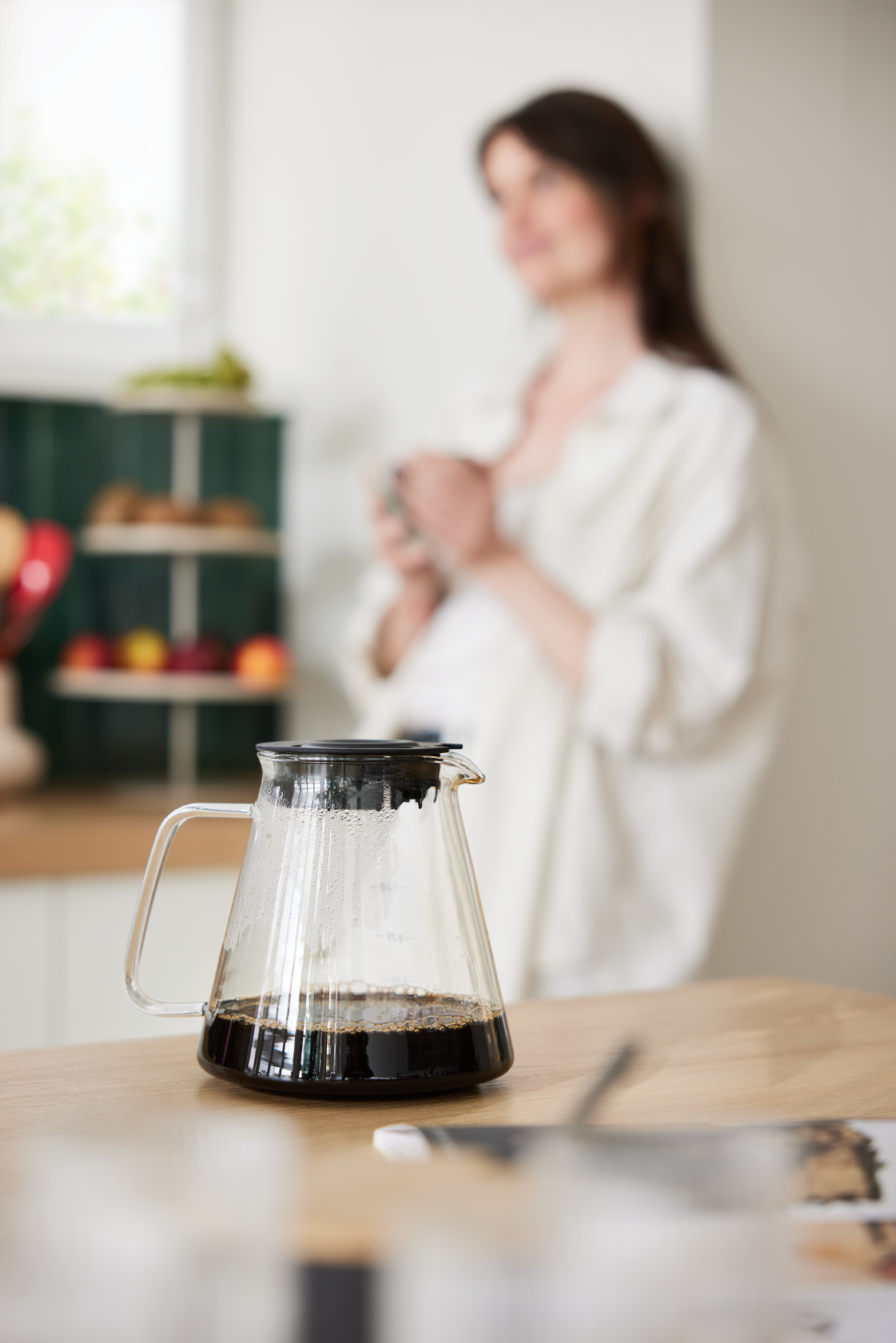 Glaskande med friskbrygget kaffe i et lyst køkken med en person i baggrunden