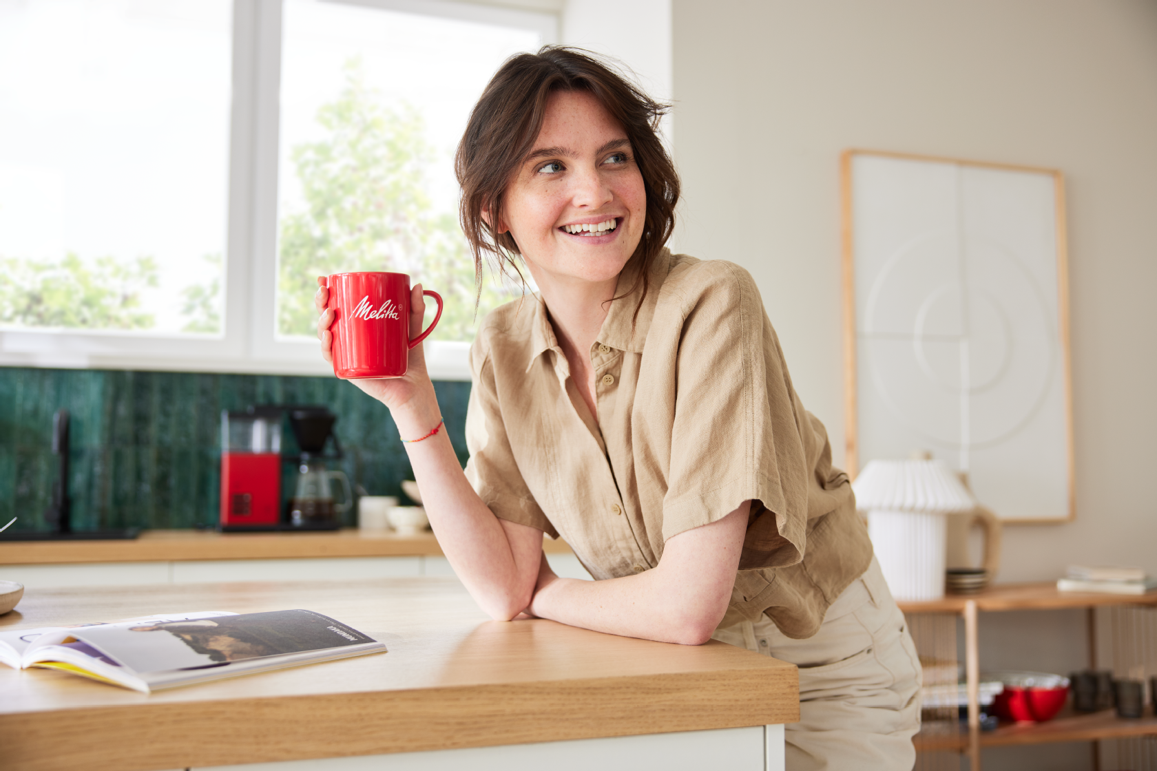 Kvinde nyder kaffe fra et rødt Melitta-krus i et moderne køkken med en Melitta ONE® filterkaffemaskine