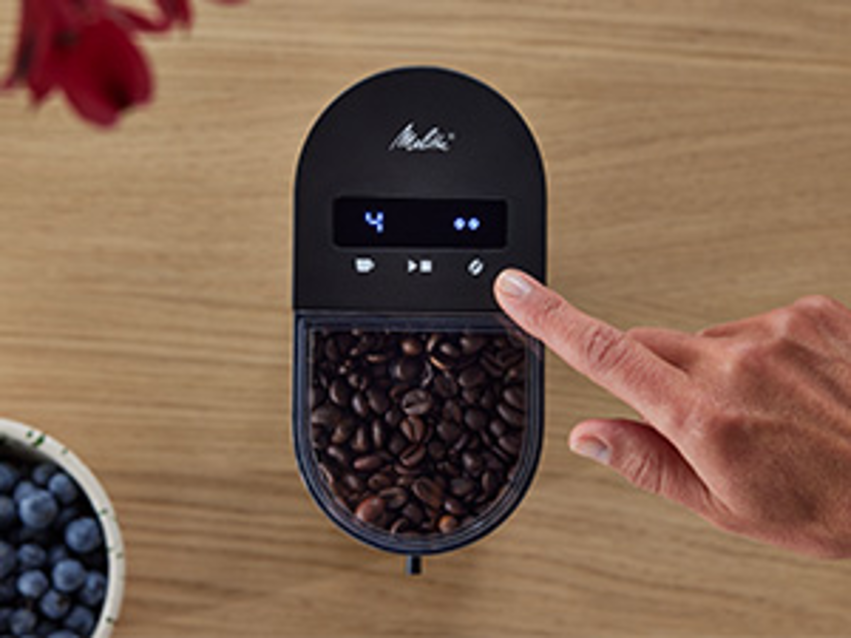 Hand bedient das digitale Bedienfeld der Melitta Linea® Kaffeemühle mit Bohnenbehälter in Draufsicht.