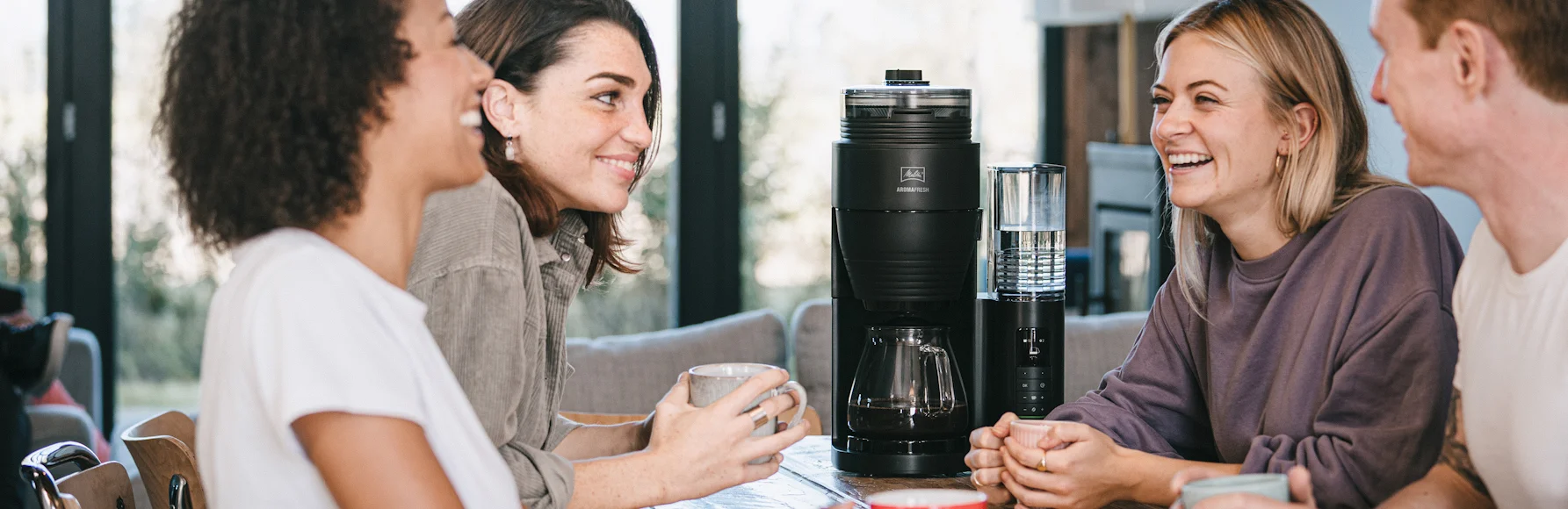 Vier mensen zitten samen met koffiemokken naast Melitta AromaFresh Pro filterkoffiezetapparaat