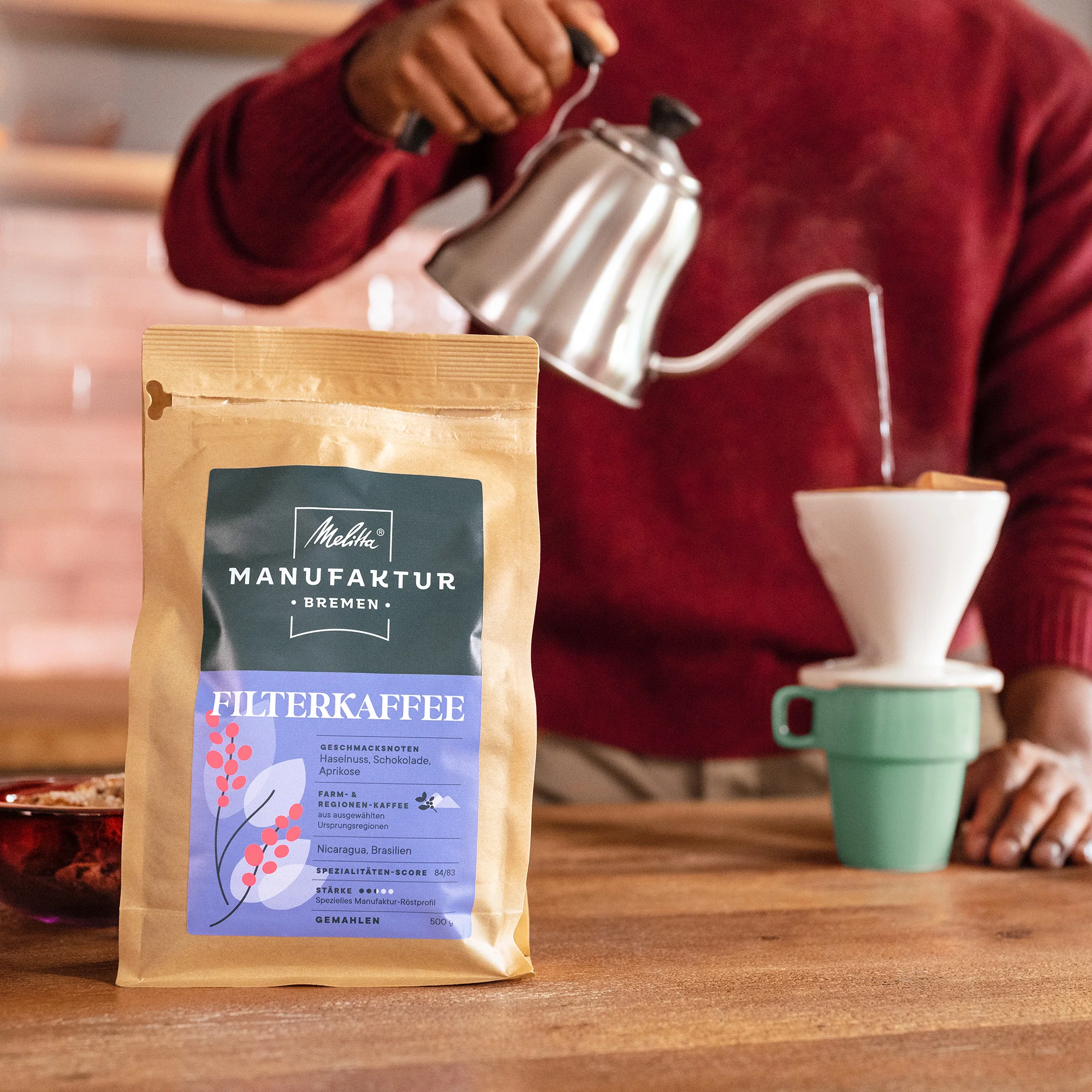 Mann gießt heißes Wasser aus Wasserkessel über einen Handfilter in eine Tasse. Daneben steht eine Packung Melitta Manufaktur Filterkaffee