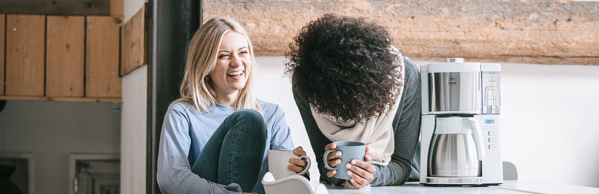 Dos mujeres disfrutan de un café junto a una cafetera de filtro Melitta Look de acero inoxidable