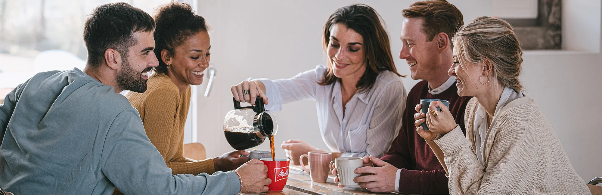 Un grupo de jóvenes está sentado alrededor de una mesa disfrutando juntos de un café Melitta recién hecho