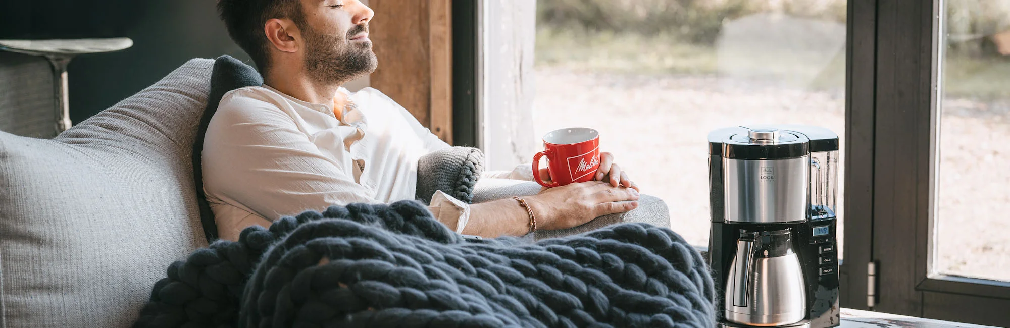 Image of a man comfortably sitting on a sofa covered with a blanket and holding a red Melitta mug in his hand.