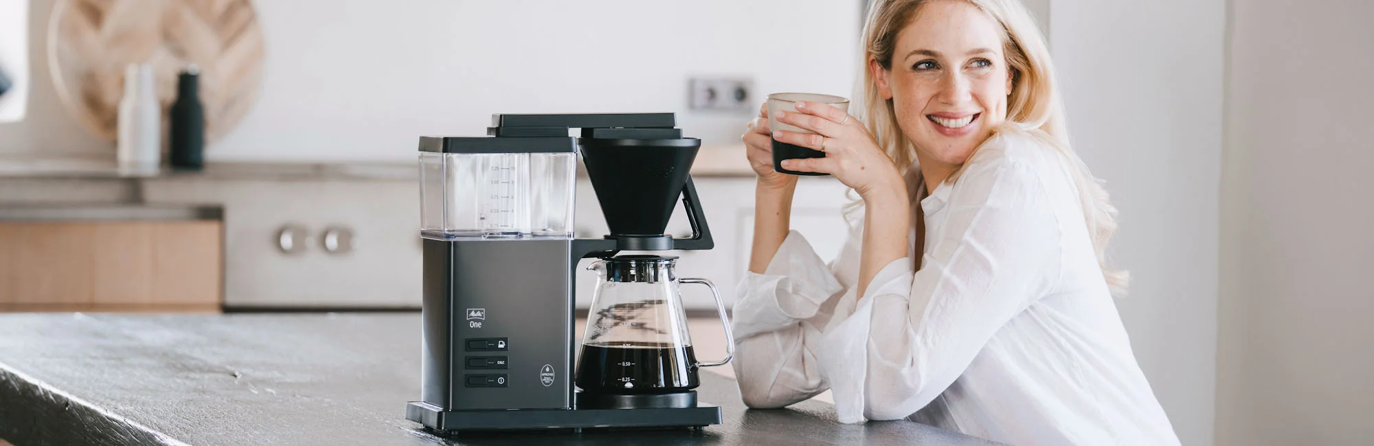Image of a woman in a kitchen drinking coffee from a coffee maker from our range of electric coffee makers.