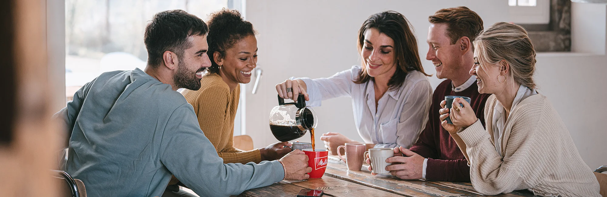 Freundesgruppe beim Kaffeegenuss, während jemand Kaffee aus einer Melitta Kanne in eine rote Melitta Tasse gießt