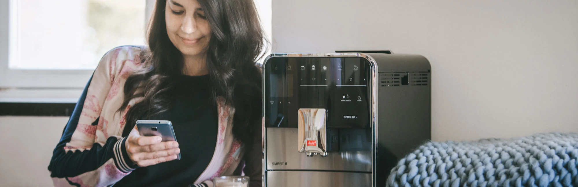 Photo of a woman choosing her coffee preparation using her mobile phone and our Barista connected automatic machine