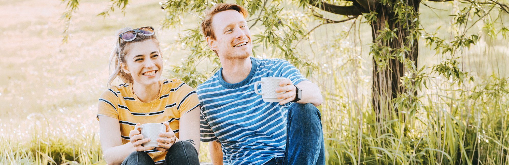 Frau und Mann sitzen draußen vor hohem Gras und Baum, lachen und haben eine Tasse Kaffee in der Hand.