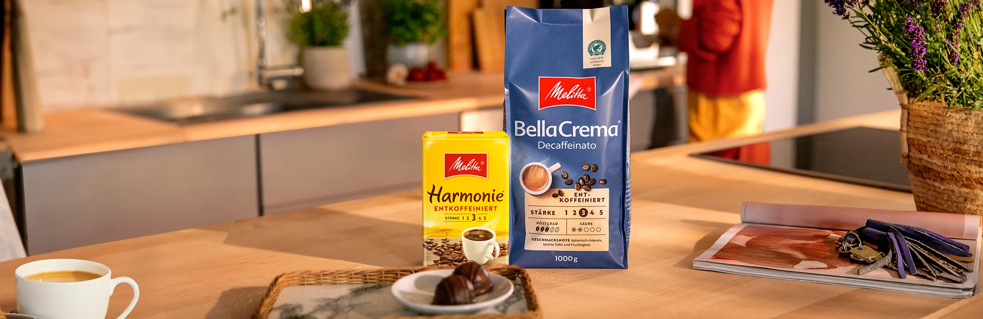 Eine Packung Melitta BellaCrema Decaf und Melitta Harmonie Decaf auf einer hölzernen Küchentheke mit einer Tasse Kaffee und Kuchen daneben