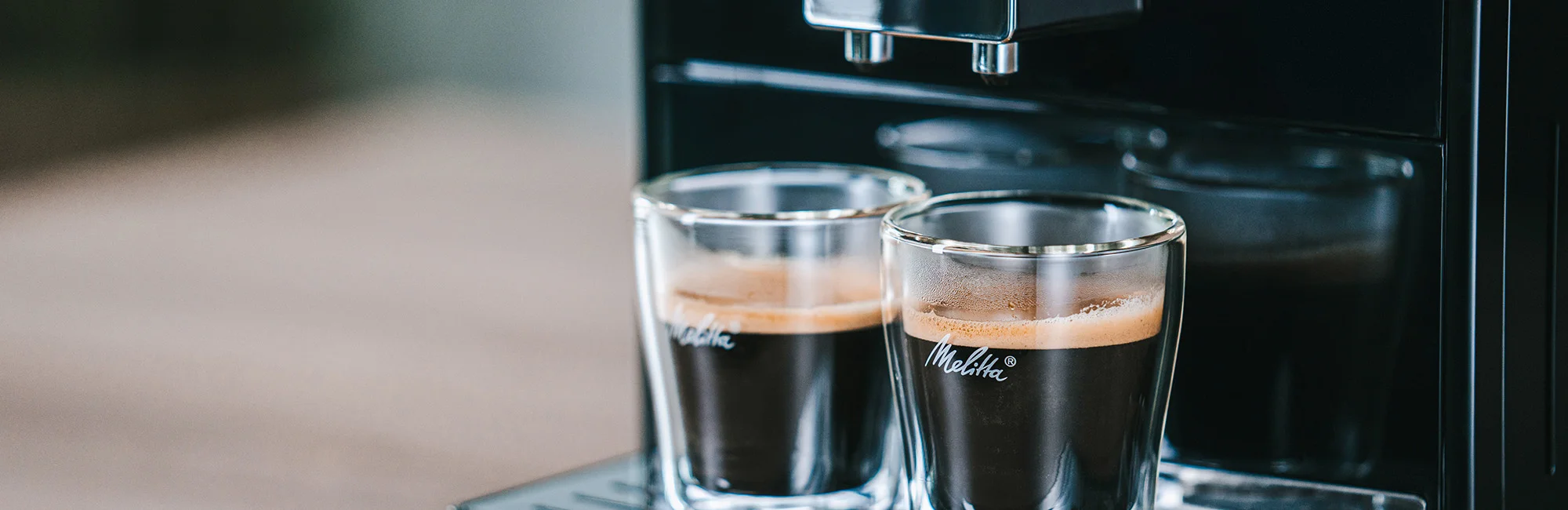 Zwei Espressi in doppelwandigen Gläsern unter einem Melitta Kaffeevollautomaten in Edelstahloptik