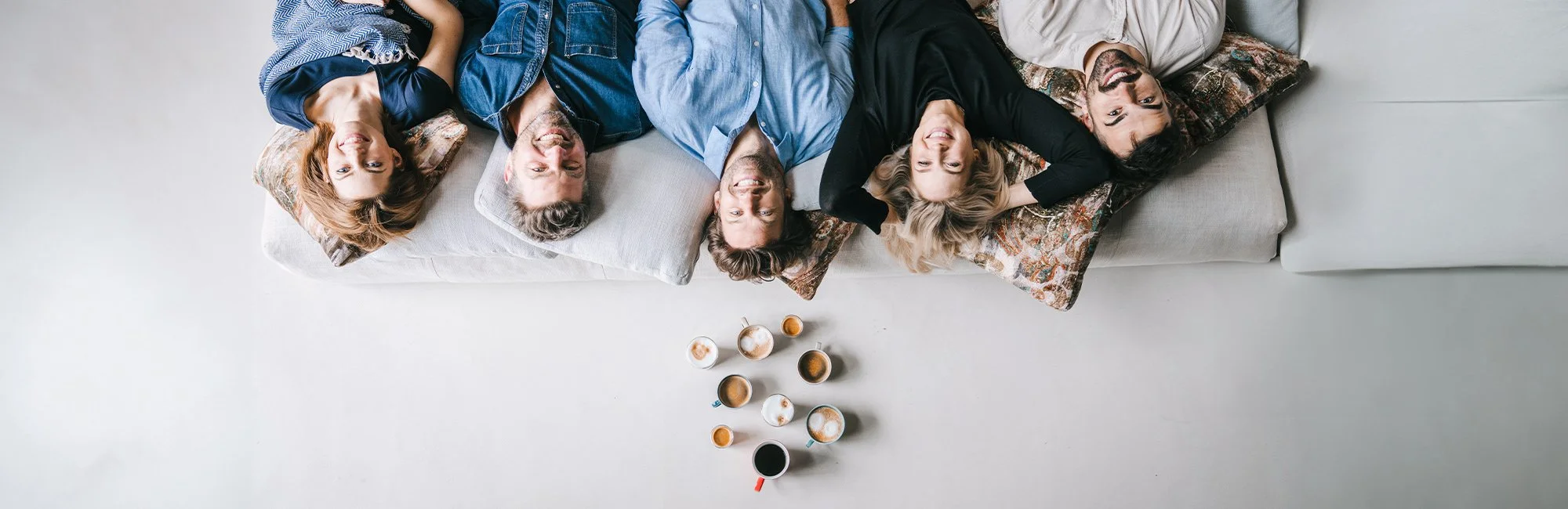 Freunde liegen lachend auf der Couch, über ihren Köpfen verschiedene Tassen mit Kaffee
