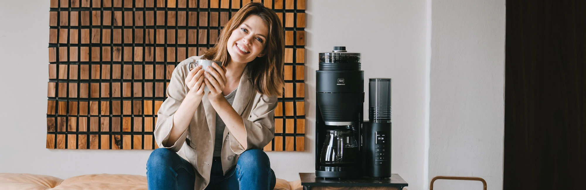 Picture of a woman sitting on a sofa. Next to her, there is a small table on which our beautiful Aromafresh electric coffee maker stands.