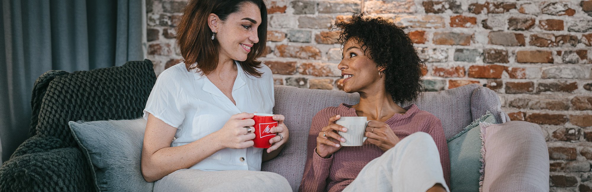 Zwei Frauen sitzen auf der Couch, lachen sich an und haben eine Tasse Kaffee in der Hand.