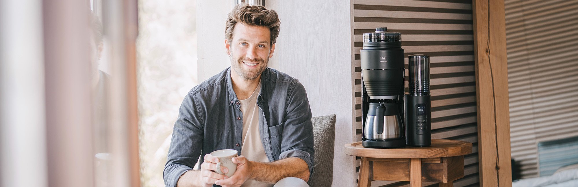 Mann mit Kaffeetasse neben schwarzer Melitta AromaFresh Filterkaffeemaschine mit Thermokanne auf einem Holztisch