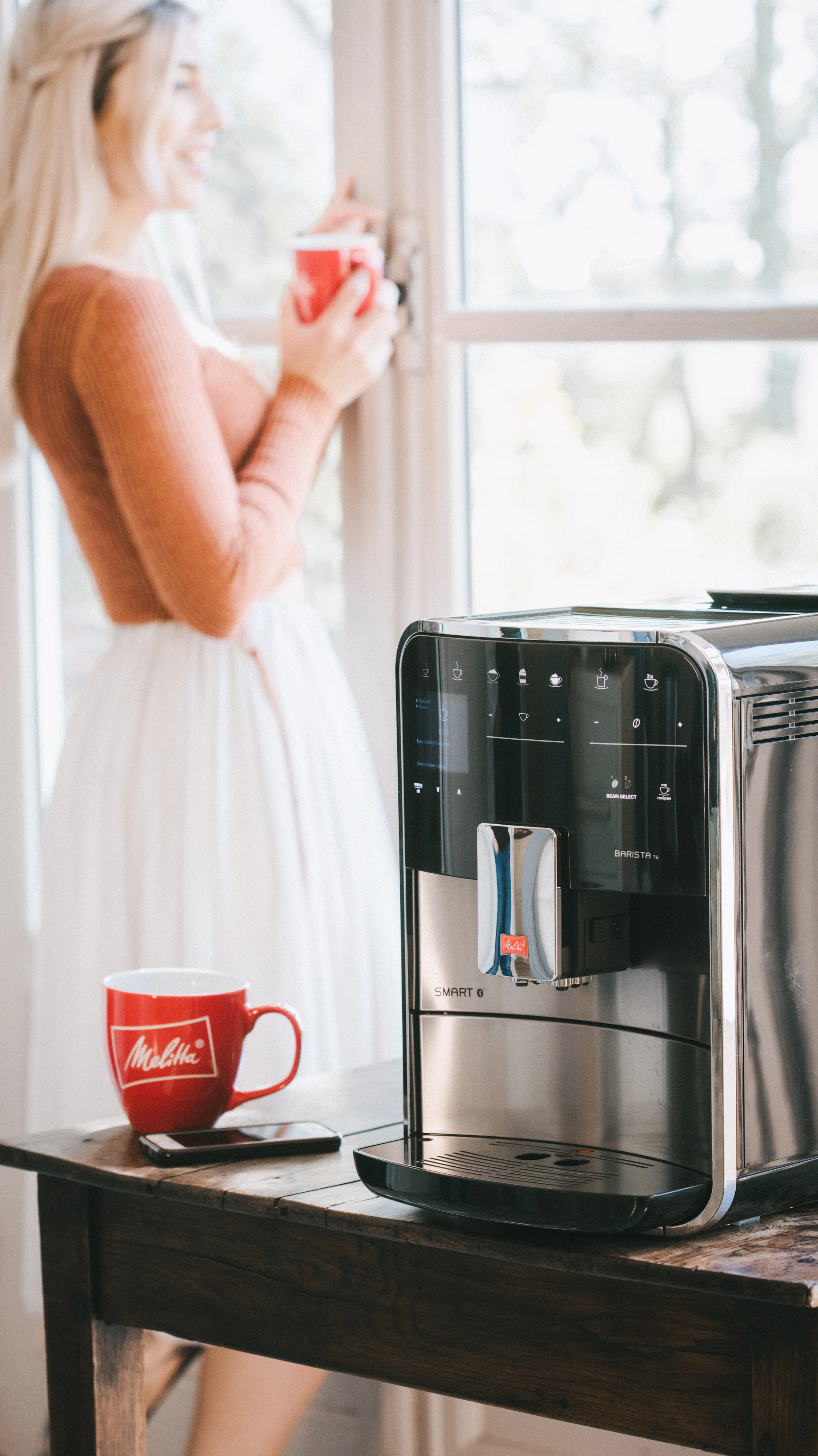 Image of a smiling woman looking at the weather outside with a cup of delicious Melitta coffee in hand.