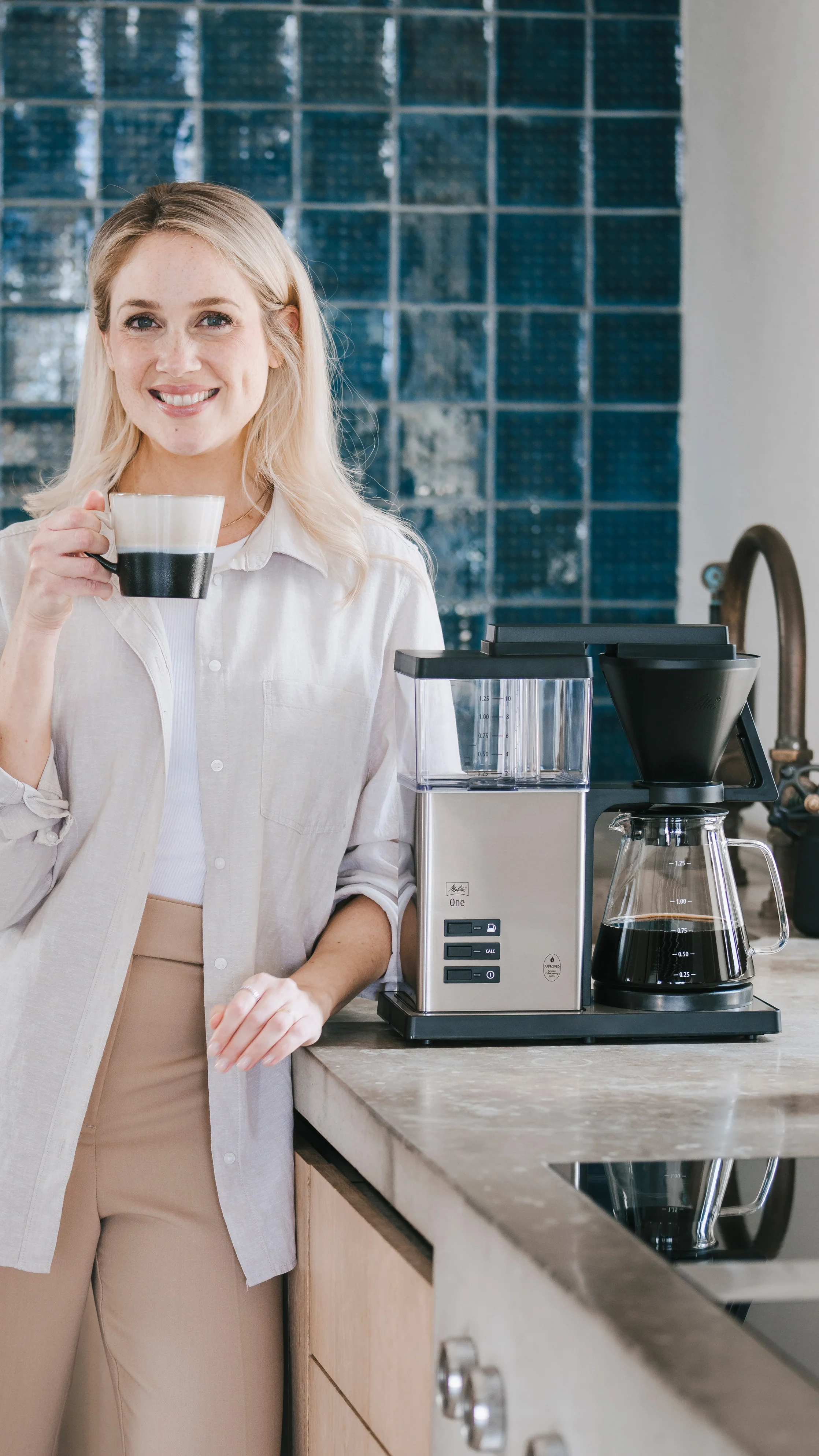 Lächelnde Frau mit Kaffeetasse neben der Melitta One aus Edelstahl.