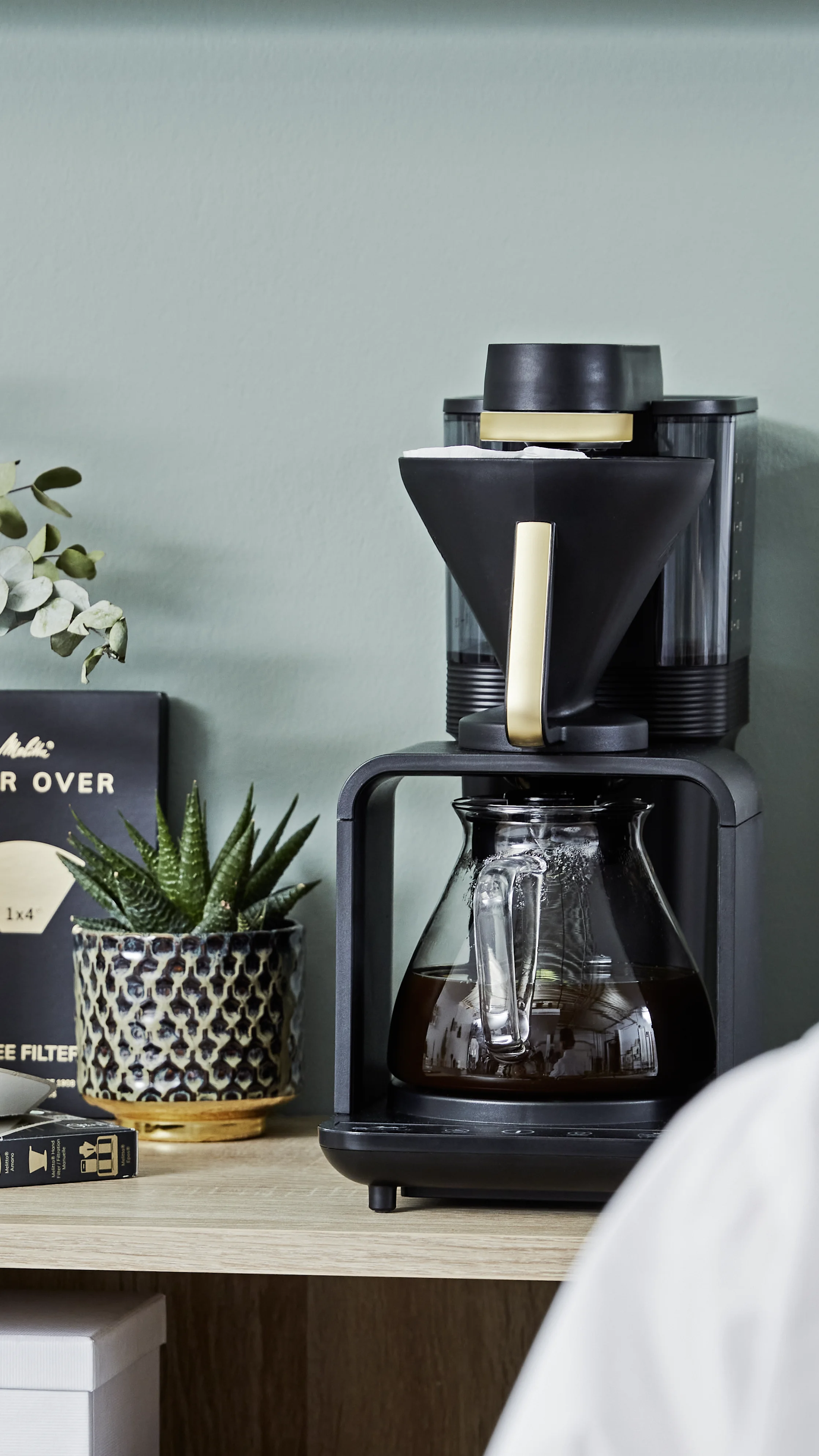 Melitta epour Filterkaffeemaschine in Schwarz mit gefüllter Glaskanne auf einem Regal neben einer Pflanze