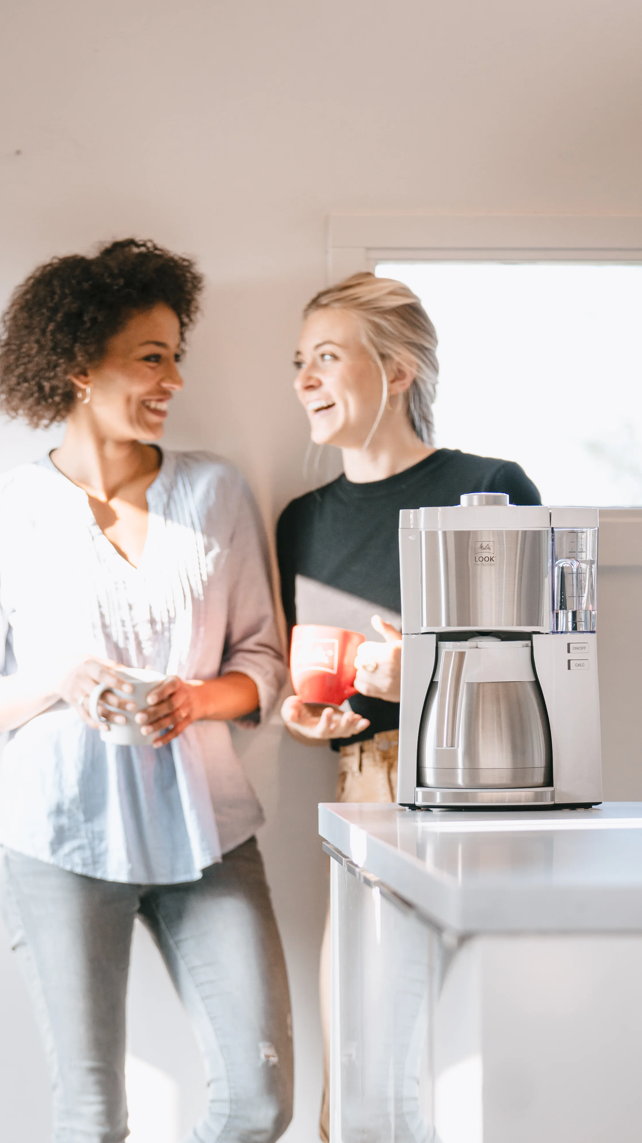 Zwei Frauen mit Kaffeetassen im Gespräch neben einer Melitta Look Therm Perfection Filterkaffeemaschine