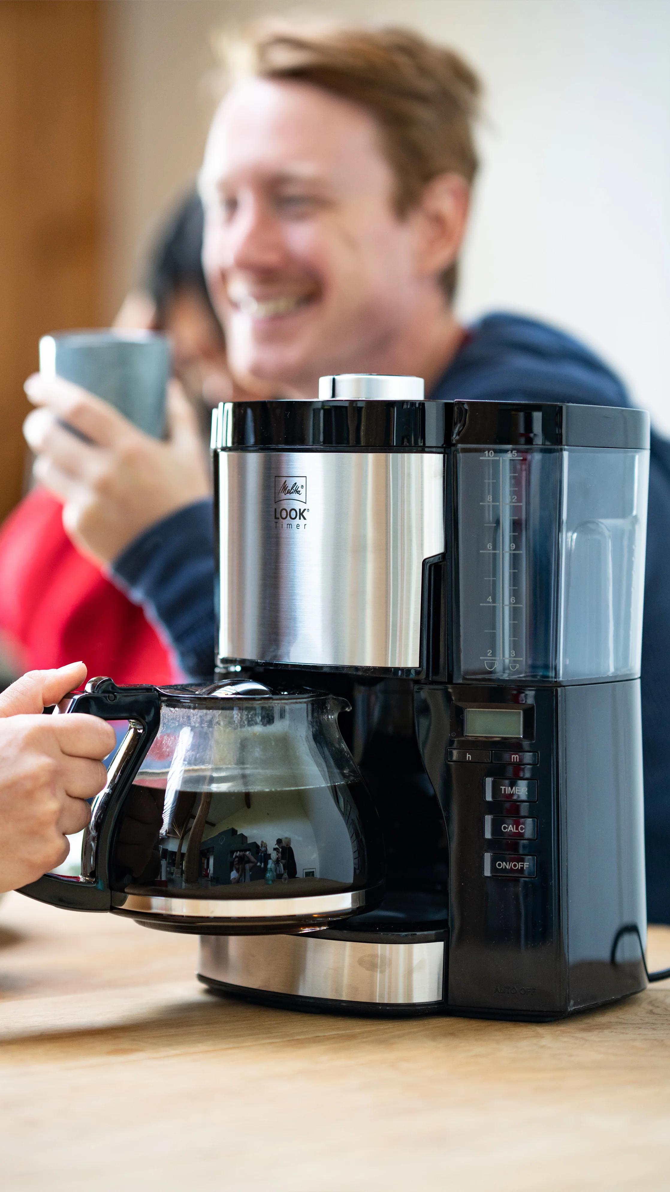 Melitta Look Timer mit Glaskanne auf Tisch, lachende Personen im Hintergrund.