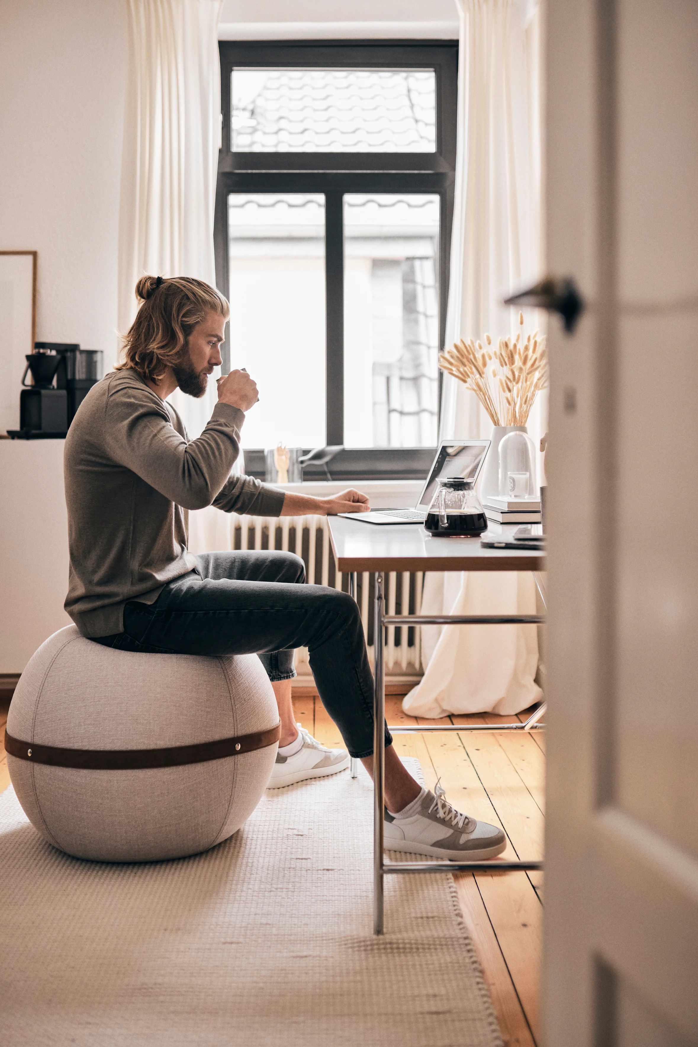 Mann sitzt am Schreibtisch an einem Laptop und trinkt aus einer Tasse.