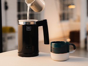 Mælk hældes i Melitta® Cremio®, ved siden af et krus med mælkeskum