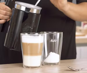 Image of a creamer pouring milk foam into 2 double-walled glasses.