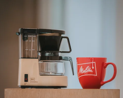 Cafetera de filtro compacta Melitta Aromaboy en beige junto a una taza roja de Melitta