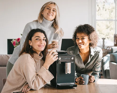 Three women are sitting at a table next to the Melitta Latticia OT fully automatic coffee machine.