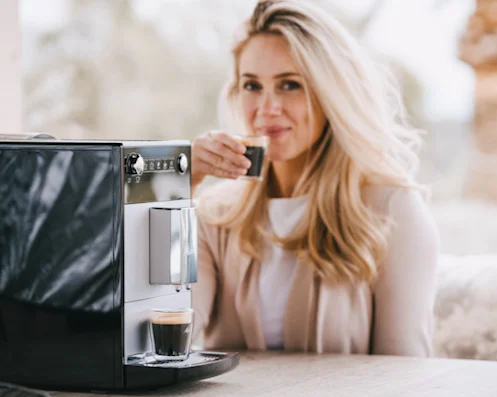A woman is sitting next to the Melitta Purista fully automatic coffee machine and drinking her espresso.