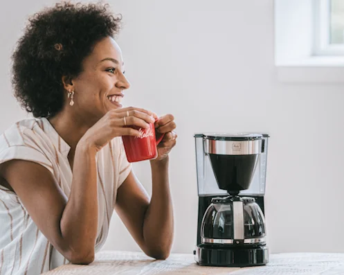 Lächelnde Frau mit roter Melitta-Tasse neben einer Melitta Filterkaffeemaschine mit Glaskanne