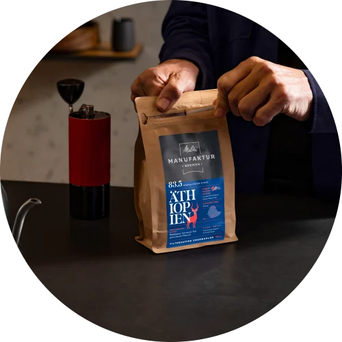 Hände, die eine Melitta Manufaktur Kaffeepackung auf einer schwarzen Küchentheke öffnen