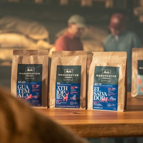 Bild aus der Melitta-Manufaktur: Kaffeepackungen stehen nebeneinander auf einem Tisch. Im Hintergrund verschwommen zwei Mitarbeiter
