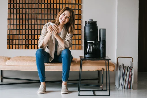 Lächelnde Frau mit Kaffeetasse sitzt auf einer Bank neben einer Melitta AromaFresh Filterkaffeemaschine