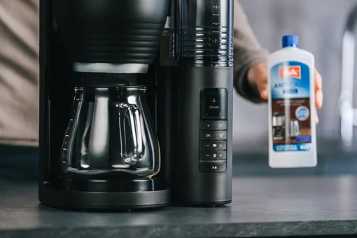 Melitta AromaFresh X Kaffeemaschine mit Glaskanne und einer Hand, die eine Flasche Melitta Anti Calc Entkalker hält.