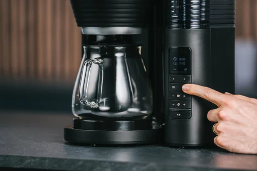 Hand drückt die OK-Taste am Bedienfeld der schwarzen Melitta AromaFresh X Kaffeemaschine mit Glaskanne.