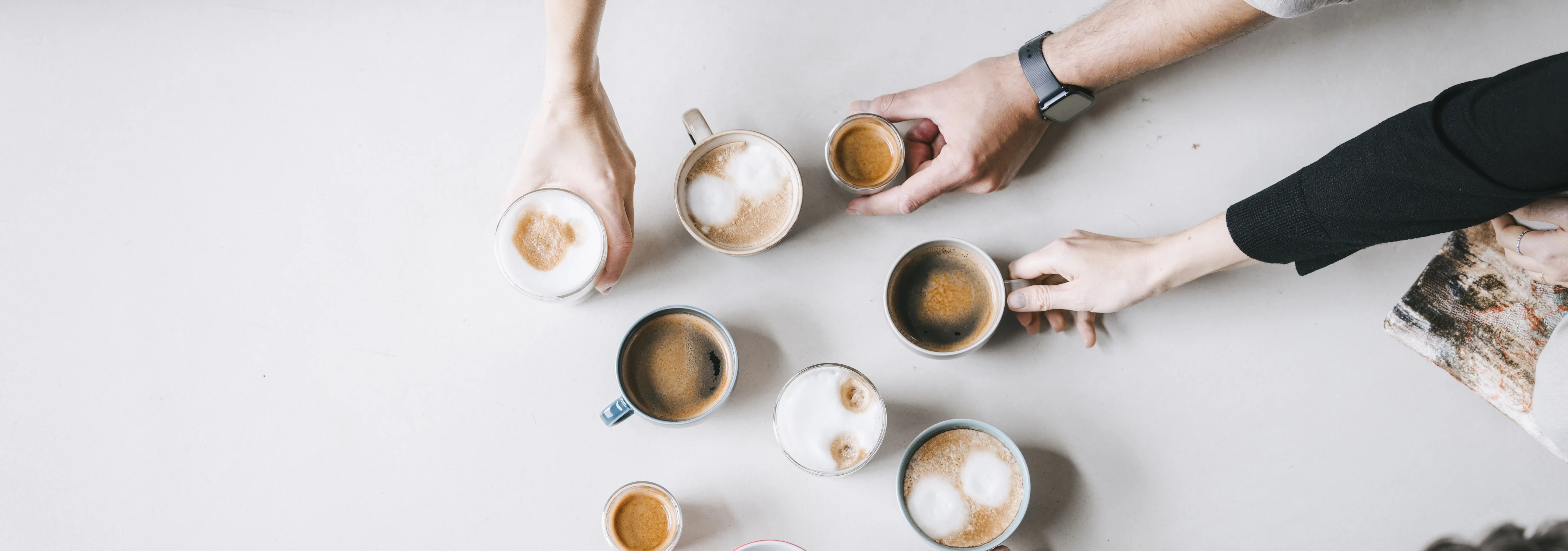 Kaffeetassen stehen auf hellem Grund, drei Hände greifen nach einer Tasse. 