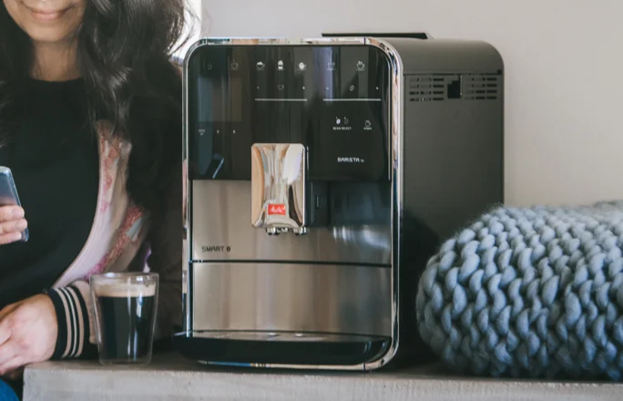une dame boit une tasse de café à côté d'une machine à expresso entièrement automatique Melitta Barista tout en vérifiant son téléphone