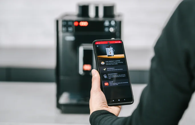 A hand holds a mobile phone in front of a Melitta Passione fully automatic machine.