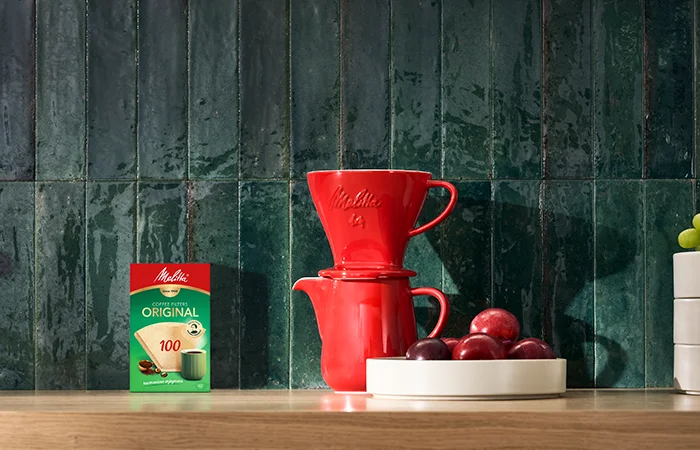 Afbeelding van een koffiepot met rode porseleinen filterhouder en één van onze dozen met 100 koffiefilters op een houten tafel.