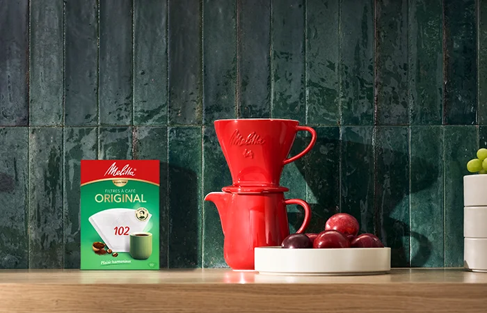 Image d'une verseuse et de son support à filtres en porcelaine rouge et d'une de nos boites de filtres à café 102 de 80 unités posées sur une table en bois