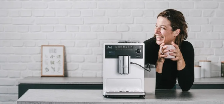 Lächelnde Frau mit Kaffeetasse neben einer silbernen Melitta LatteSelect in moderner Küche