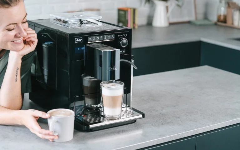 Frau genießt Kaffee neben einer schwarzen Melitta CI Touch, die einen Latte Macchiato in moderner Küche zubereitet
