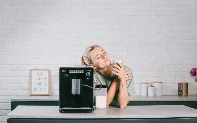 Glückliche Frau genießt einen Latte Macchiato neben einer schwarzen Melitta LatteSelect in moderner Küche