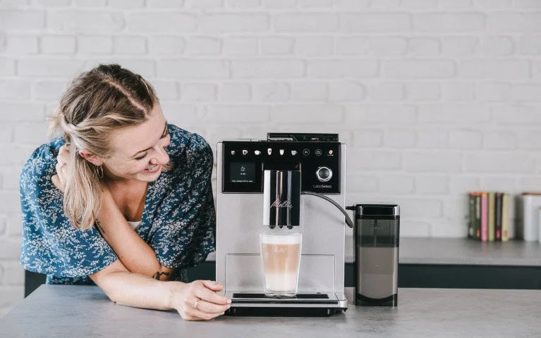 Lächelnde Frau schaut zu, wie die silberne Melitta LatteSelect einen Latte Macchiato in moderner Küche zubereitet