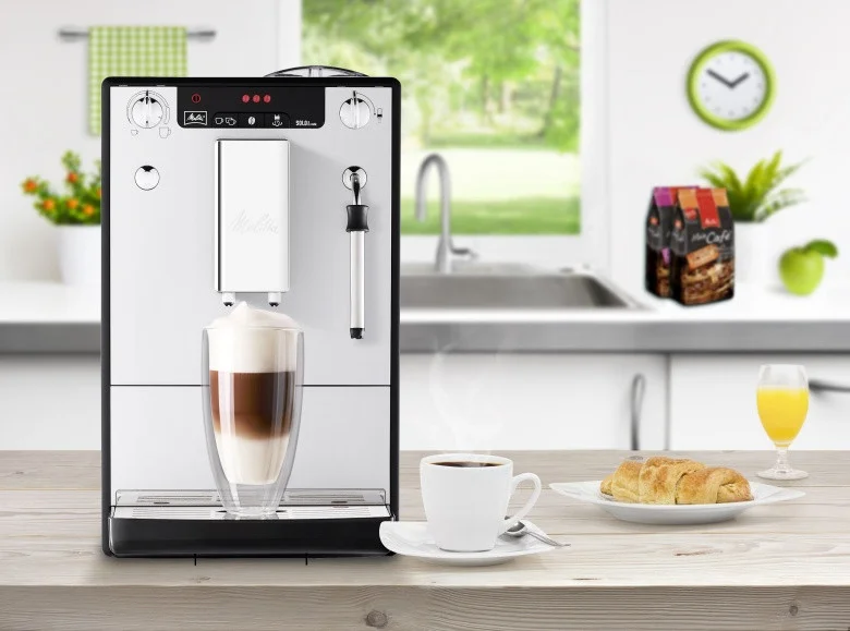 Silberne Melitta Caffeo Solo & Milk bereitet einen Latte Macchiato zu, mit Kaffeetasse, Croissant und Saft in heller Küche