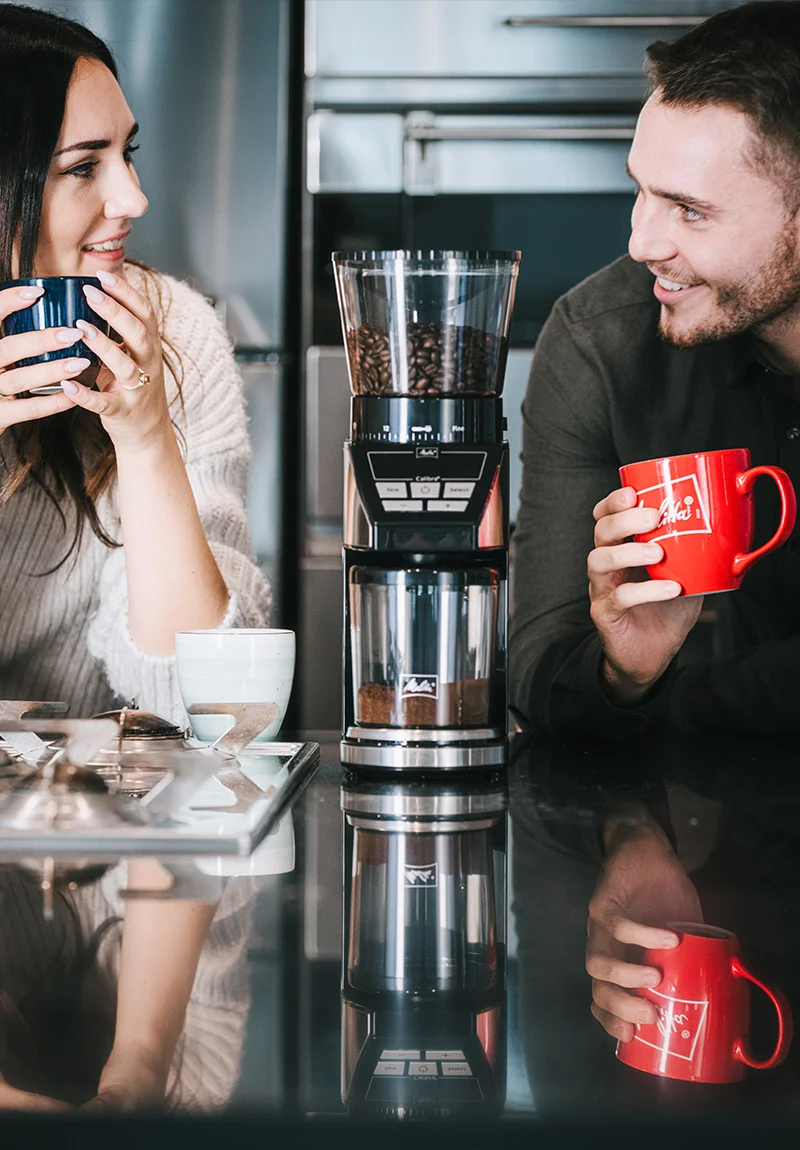 Zwei Menschen trinken Melitta Kaffee, im Vordergrund steht die Melitta Calibra Kaffeemühle.